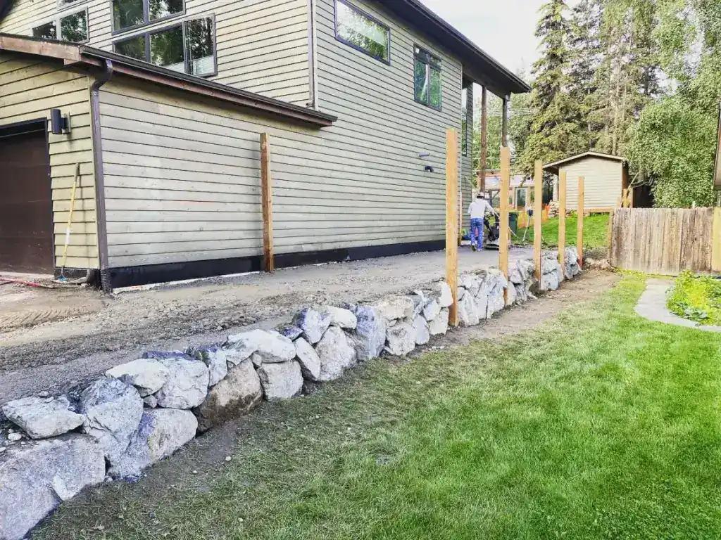 Boulder retaining wall built along a house with new fence posts set behind it